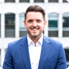 Outdoor headshot of Edd Micklem smiling directly at the camera wearing a blue suit jacket and shirt with the cityscape in the background.