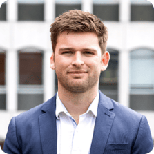 Outdoor headshot of Will Goodwin smiling directly at the camera wearing a blue suit jacket and shirt with the cityscape in the background.